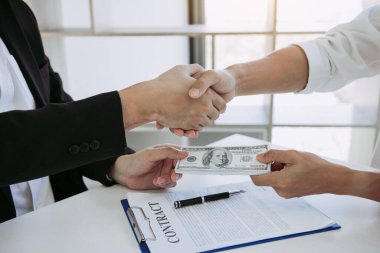 Two corporate businessmen shaking hands while one man places money on document in office room with corruption concept. clipart