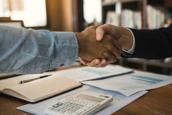 Entrepreneurs collaboration deal shaking hands in a modern office and financial paper graph on desk.