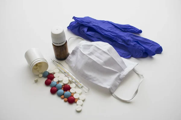 Beyaz arka planda cerrahi eldivenler, bir maske, bir termometre ve iki kavanoz hap var..