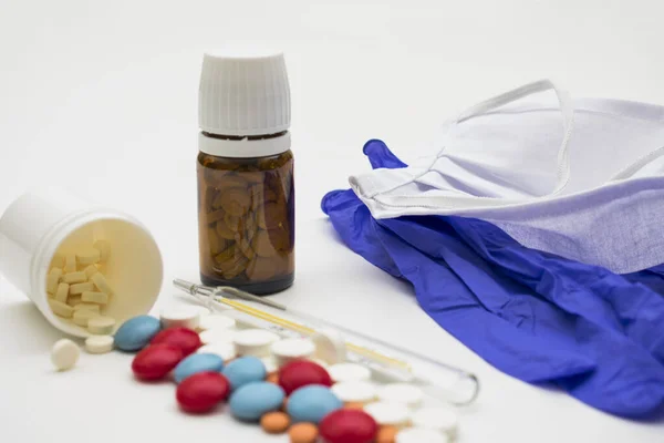 Beyaz arka planda cerrahi eldivenler, bir maske, bir termometre ve iki kavanoz hap var..