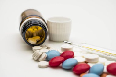 Beyaz arka planda haplar, ters çevrilmiş bir kavanoz hap ve bir termometre var..