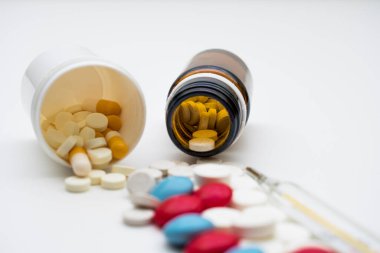 Beyaz arka planda haplar, ters çevrilmiş bir kavanoz hap ve bir termometre var..