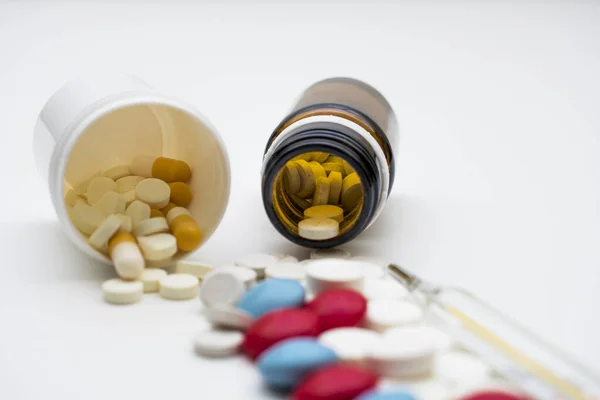 Beyaz arka planda haplar, ters çevrilmiş bir kavanoz hap ve bir termometre var..