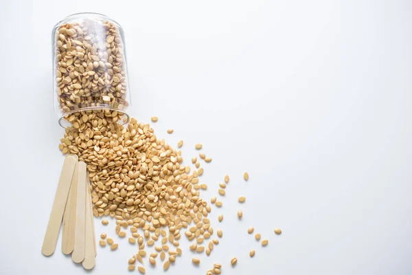 Altın balmumu granülleri ve beyaz arka planda izole edilmiş tahta çubukları olan cam bir kavanoz.. 