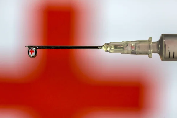 Hypodermic needle dripping fluid with red cross reflected over a red and white out of focus background