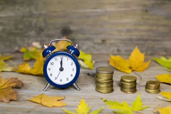 Alarm clock and money coins on wooden rustic old table with autumn leaves. Time is money autumn style comcept. Discount time concept. Time for fall autumn savings. Time to make money.