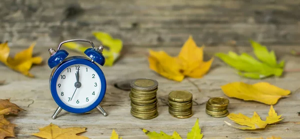 Alarm clock and money coins on wooden rustic old table with autumn leaves. Time is money autumn style comcept. Discount time concept. Time for fall autumn savings. Time to make money.