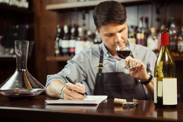 Masculino sommelier degustação de vinho tinto e fazer notas no balcão de bar — Fotografia de Stock