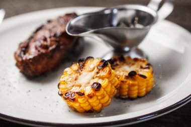 Biftek ızgara mısır ve beyaz plaka üzerinde demir kapta sos ile