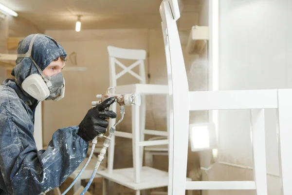 Homem que pinta a cadeira na pintura branca na máscara respiratória. Aplicação de retardador de chama garantindo proteção contra incêndio, pulverização airless . — Fotografia de Stock