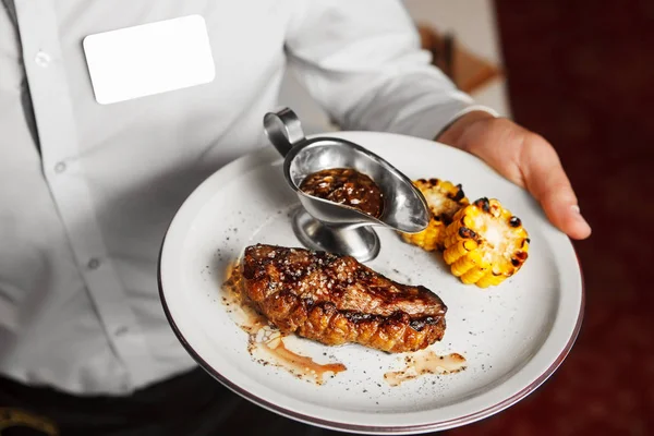 Waiter serve on plate beefsteak with grilled corn and sauce