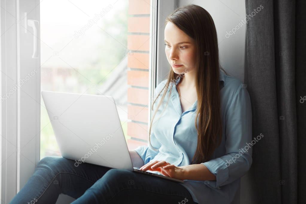 Young woman works at laptop and sits at windowsill