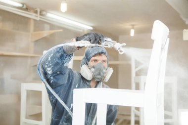 Man painting chair into white paint in respiratory mask. Application of flame retardant ensuring fire protection, airless spraying. clipart