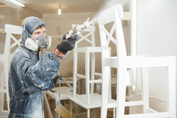 Hombre pintando silla en pintura blanca en máscara respiratoria. Aplicación de retardante de llama que garantiza la protección contra incendios, pulverización sin aire . — Foto de Stock