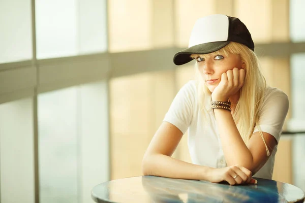 테이블에 앉아 하는 금발 머리를 가진 여자. 젊은 여자 야구 모자와 트림된 앞 머리. — 스톡 사진