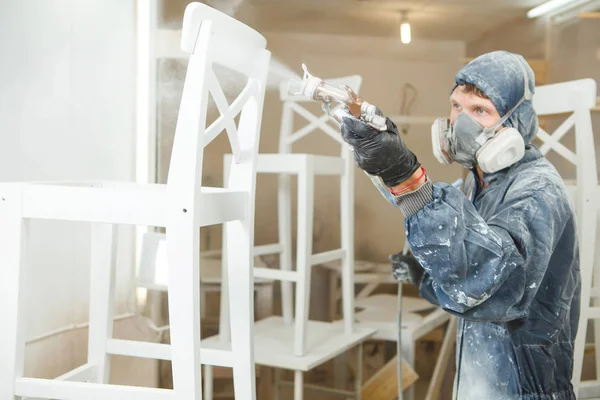 Homem que pinta a cadeira na pintura branca na máscara respiratória. Aplicação de retardador de chama garantindo proteção contra incêndio, pulverização airless . — Fotografia de Stock