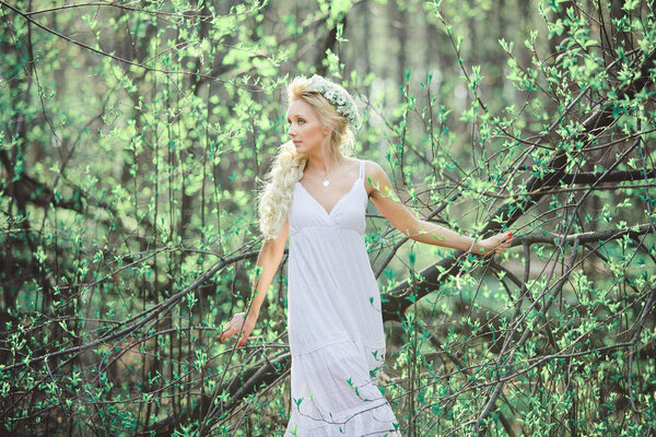 Beautiful woman in white dress and floral wreath on her head among the blossoming leaves. Portrait of a cute blond girl in the woods.