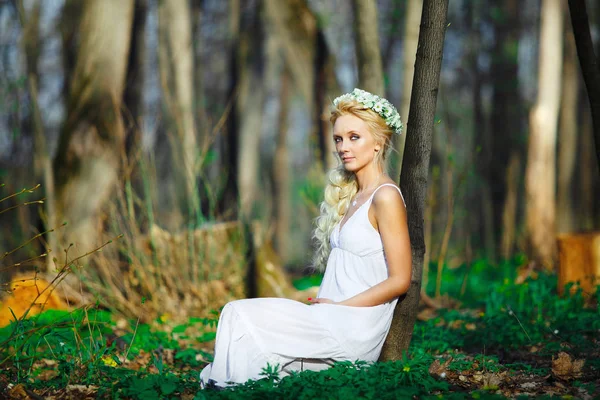 Bella donna in abito bianco con bella ghirlanda sulla testa siede da albero foresta verde . — Foto Stock