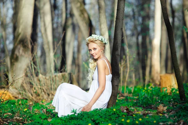 Bella donna in abito bianco con bella ghirlanda sulla testa siede da albero foresta verde . — Foto Stock