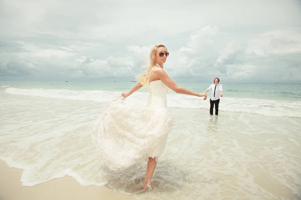 Νύφη στο γάμο φόρεμα τρέχει να γαμπρό πάνω στη θάλασσα, γυρίζει πίσω. τυχερή γυναίκα στην παραλία. — Φωτογραφία Αρχείου