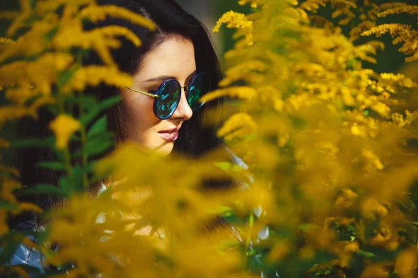 Mujer étnica caucásica con pelo negro lleva gafas con estilo entre flores amarillas . — Foto de Stock