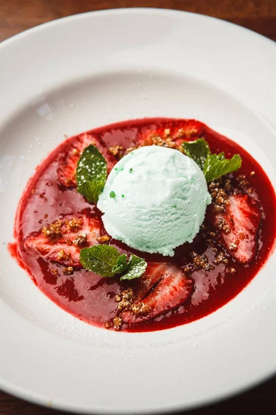 turquoise mint ice cream on plate with addition sauce, pieces of strawberries and mint leaves.