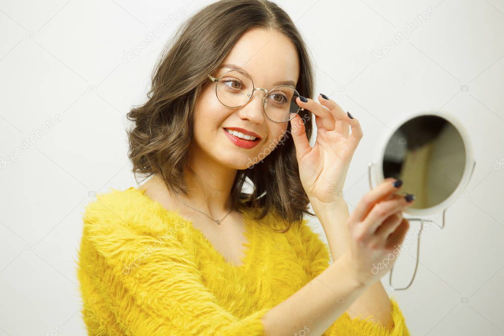 horizontal portrait of woman picks up round stylish spectacles and admires the mirror over white isolated background.