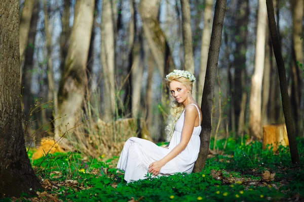 Beautiful woman in white dress with beautiful wreath on head sits by tree green forest. — Stock Photo, Image
