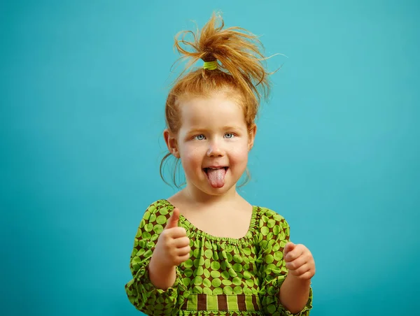 Retrato de menina de três anos de idade preso a língua para fora e mostra polegar para cima, tem cabelo vermelho, vestido com vestido verde, expressa olhar sincero, fica sobre fundo isolado azul. Criança de bom humor . — Fotografia de Stock