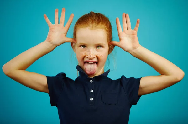 Portrait de vilaine fille aux cheveux roux avec des taches de rousseur sortir sa langue et vous taquine, en mettant les doigts à la tête, debout sur fond bleu isolé . — Photo