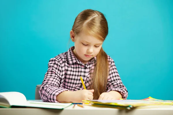 Chica se sienta en el escritorio y escribe a la libreta, sobre fondo azul aislado. Estudiante de primer grado en la escuela . — Foto de Stock