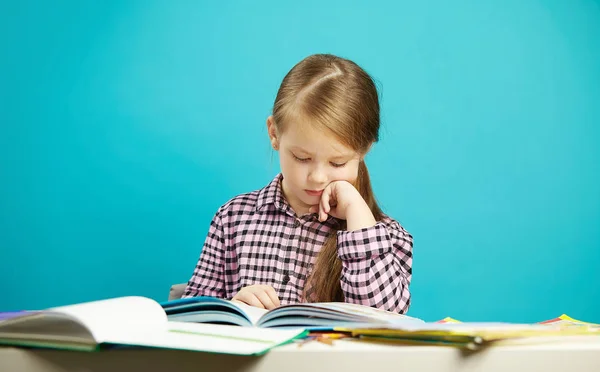 Retrato aislado de niña sobre fondo azul, sentada en el escritorio, leyendo el libro cuidadosamente con la mano en la barbilla. Niño está interesado en aprender . — Foto de Stock