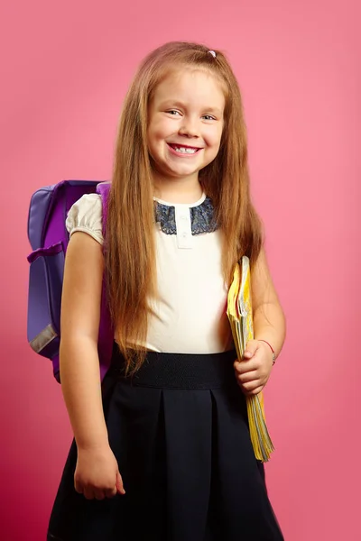 Retrato de menina sorridente em uniforme escolar com mochila no ombro e caderno em suas mãos fica em fundo rosa isolado. Estudante de bom humor vai estudar . — Fotografia de Stock