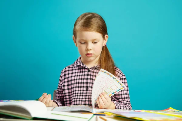 Chica se sienta en el escritorio en el aula, da vuelta a la página del libro de texto, sostiene el lápiz, sobre fondo azul aislado. El escolar en la escuela recibe conocimiento . — Foto de Stock