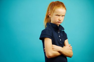 Kızgın ifade ile memnun kız izole mavi arka plan üzerinde duruyor, ifade hayal kırıklığı veya memnuniyetsizlik, gösterir kötü ruh hali, katlanmış elleriyle haç, furrowed yüz.