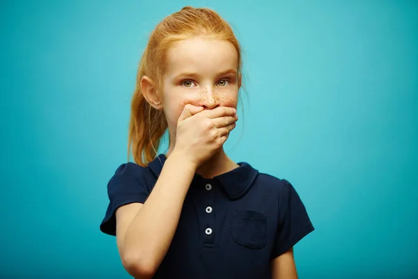 Red-haired girl covered her mouth with hand and looks at camera with concern over isolated blue background, expresses privacy, does not confess to what she did.
