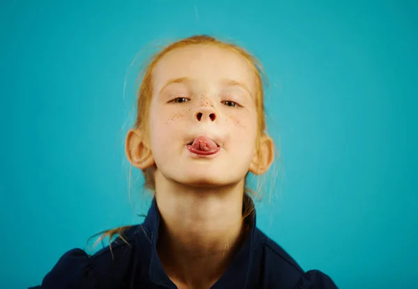 Close-up retrato de menina ruiva impertinente no fundo isolado azul mostra-lhe a língua e brinca . — Fotografia de Stock