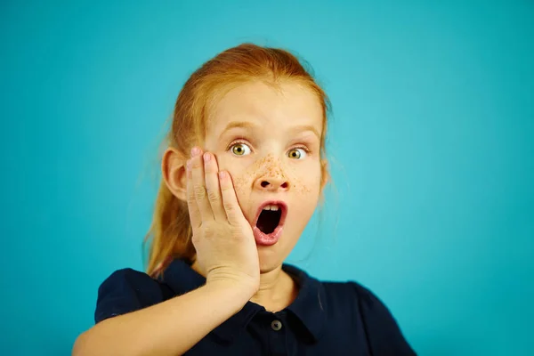 Retrato de cerca de niña pelirroja de siete años con emoción pronunciada, pone la mano en la mejilla, expresa sorpresa o asombro, demuestra confusión y miedo sobre fondo azul aislado . —  Fotos de Stock