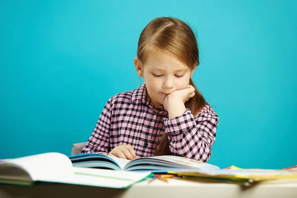 Disciplined girl attentively reads book sitting at the table, expresses her passion, on isolated blue background. Child performs home task. — Stock Photo, Image