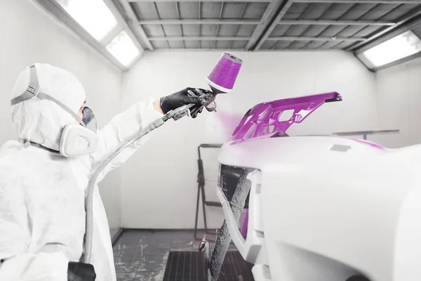 Car painter spraying violet paint with spray gun on bumper of vehicle in paint chamber.