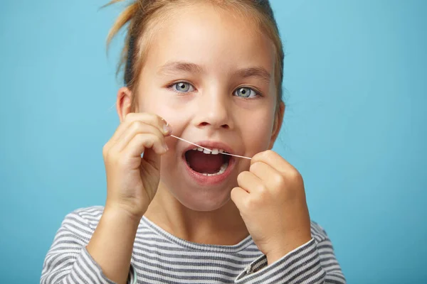 Blanc enfant fille à l'aide d'un fil dentaire sur fond bleu isolé et souriant tout en regardant la caméra . — Photo