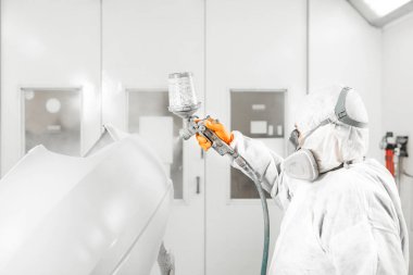 Maskeli ve koruyucu iş elbiseli bir otomobil tamircisi boya odasında beyaz araba gövdesi boyuyor..