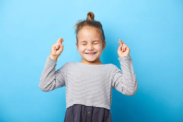 Fröhliches kleines Mädchen beißt die Zähne zusammen, drückt die Daumen, macht Wünsche, drückt die Augen zu, wartet auf gute Nachrichten, steht drinnen vor blauem Hintergrund. — Stockfoto