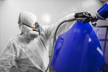 Erkek işçi arabanın gövdesine mavi boya püskürtüyor. Araba bakım servisinin boya odasında..