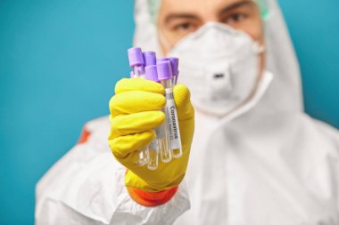 Erkek hemşire Coronavirus için tıbbi test tüpleri tutuyor. Koruyucu tulumlu ve solunum maskeli bir tıp çalışanı CV19 'dan örnek almak üzere..