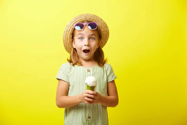 여름옷을 입은 어린 소녀의 스튜디오 초상화, 놀라움의 감정을 표현하는 것, 밀짚 모자와 선글라스를 쓰는 것, 손에 아이스크림을 들고 있는 것, 노란색외진 곳에 서 있는 것. — 스톡 사진