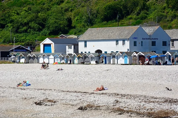 Los turistas en la playa de guijarros con cabañas de playa y la Academia de construcción de barcos en la parte trasera, Lyme Regis . — Foto de Stock