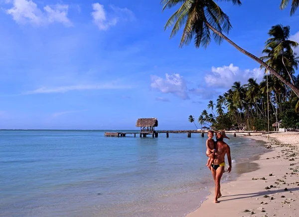 Homem caminhando ao longo da praia segurando uma criança com vista para o molhe em Pigeon Point, Tobago . — Fotografia de Stock