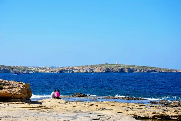 Pár sedí na skalnaté pobřeží s výhledem na zátoku, Bugibba, Malta. — Stock fotografie