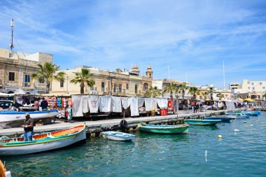 Geleneksel Malta Dghajsa balıkçı tekneleri ve liman waterfront binalar ve Pazar tezgahları ile arka, Marsaxlokk, Malta.